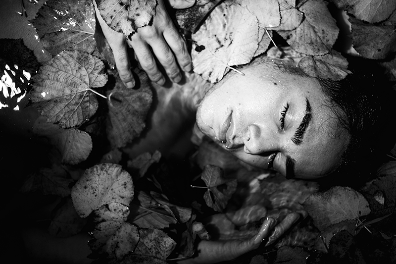 Una fotografía de bellas artes en blanco y negro, llena de alma, de Arjan Spannenburg, que presenta a una persona sumergida en agua rodeada de hojas de otoño, simbolizando el delicado equilibrio de la protección, la transparencia y el toque humano en nuestro compromiso con la privacidad.