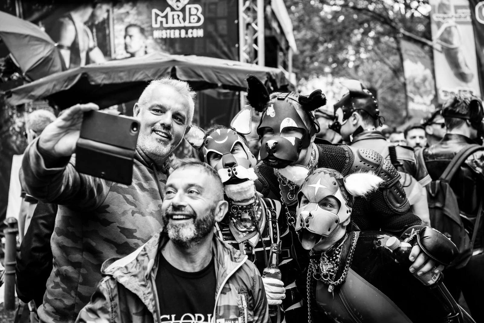 A group of people taking a selfie at an outdoor street festival, featuring several individuals wearing intricate pup-play masks and leather gear, illustrating the community aspect of fetish culture.