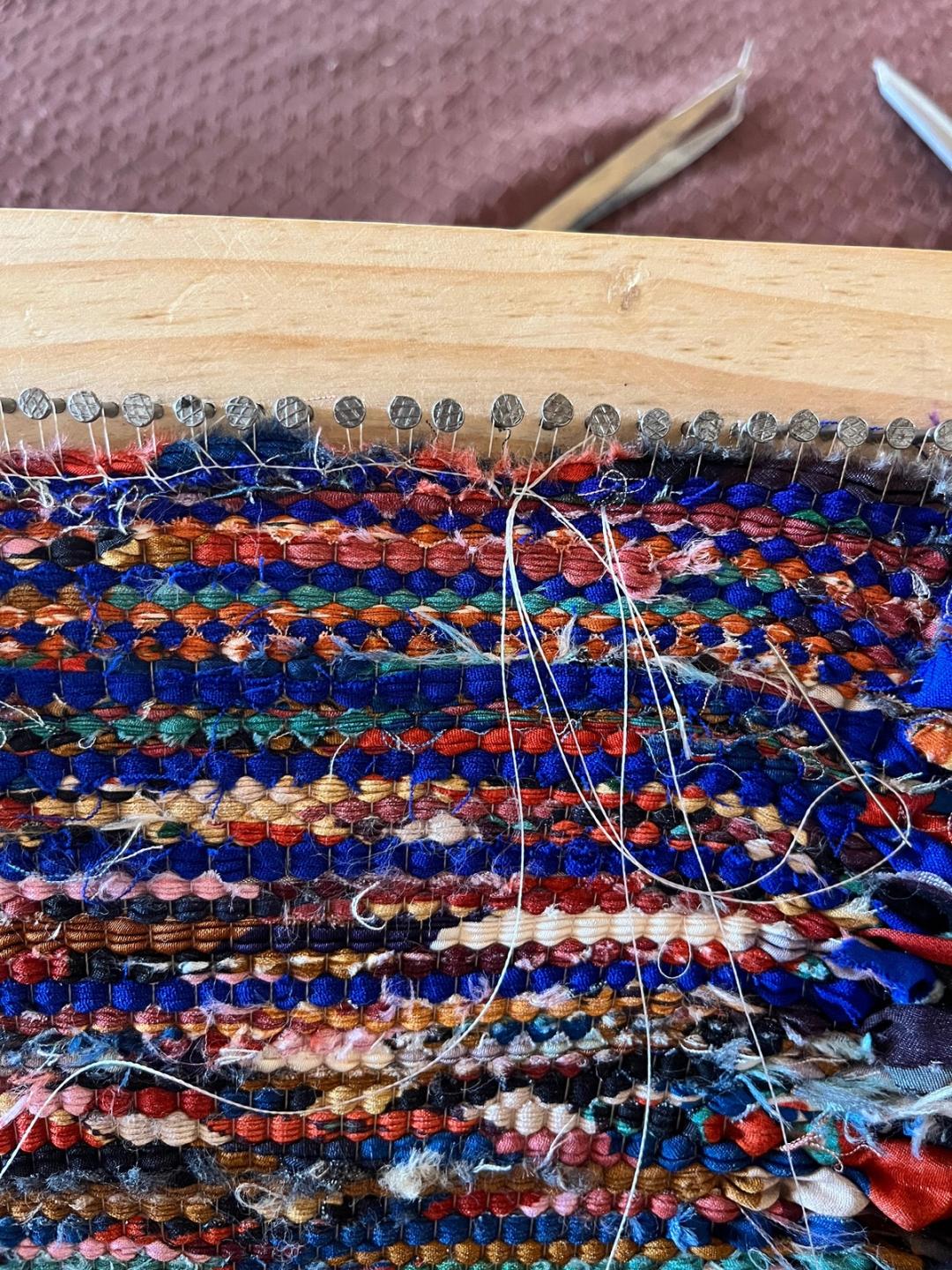 Tissage de bandes de tissus multicolores en cours sur un cadre en bois à clous, fils de chaîne apparents.
