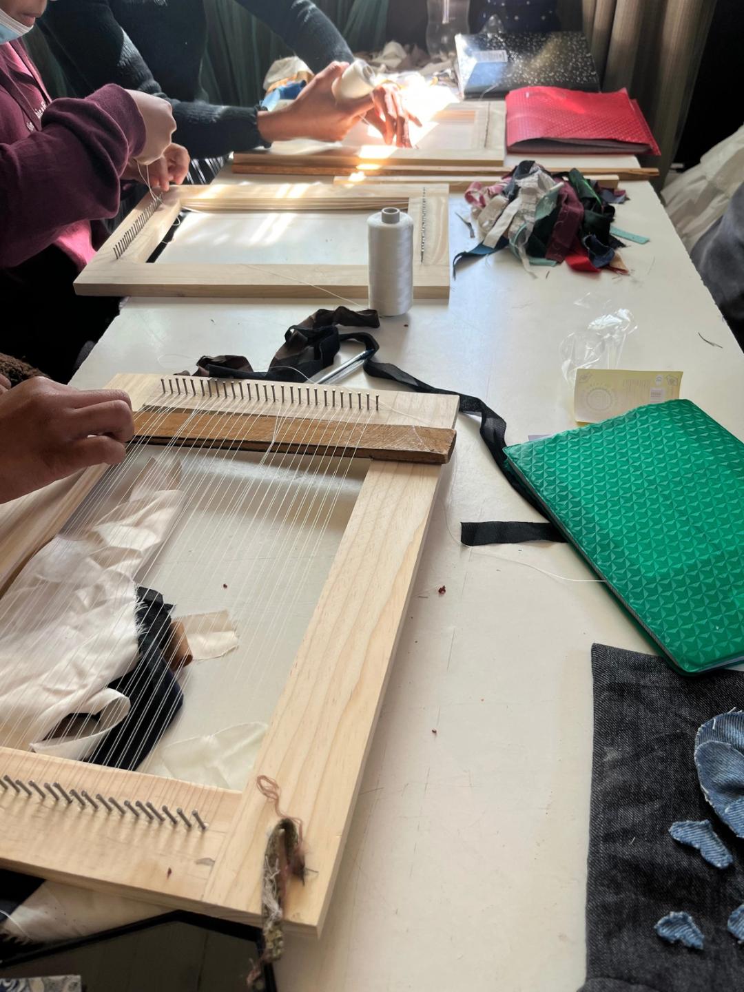 Plusieurs personnes montent des fils de chaîne sur des cadres à clous ; chutes de tissus et cône de fil sur la table.