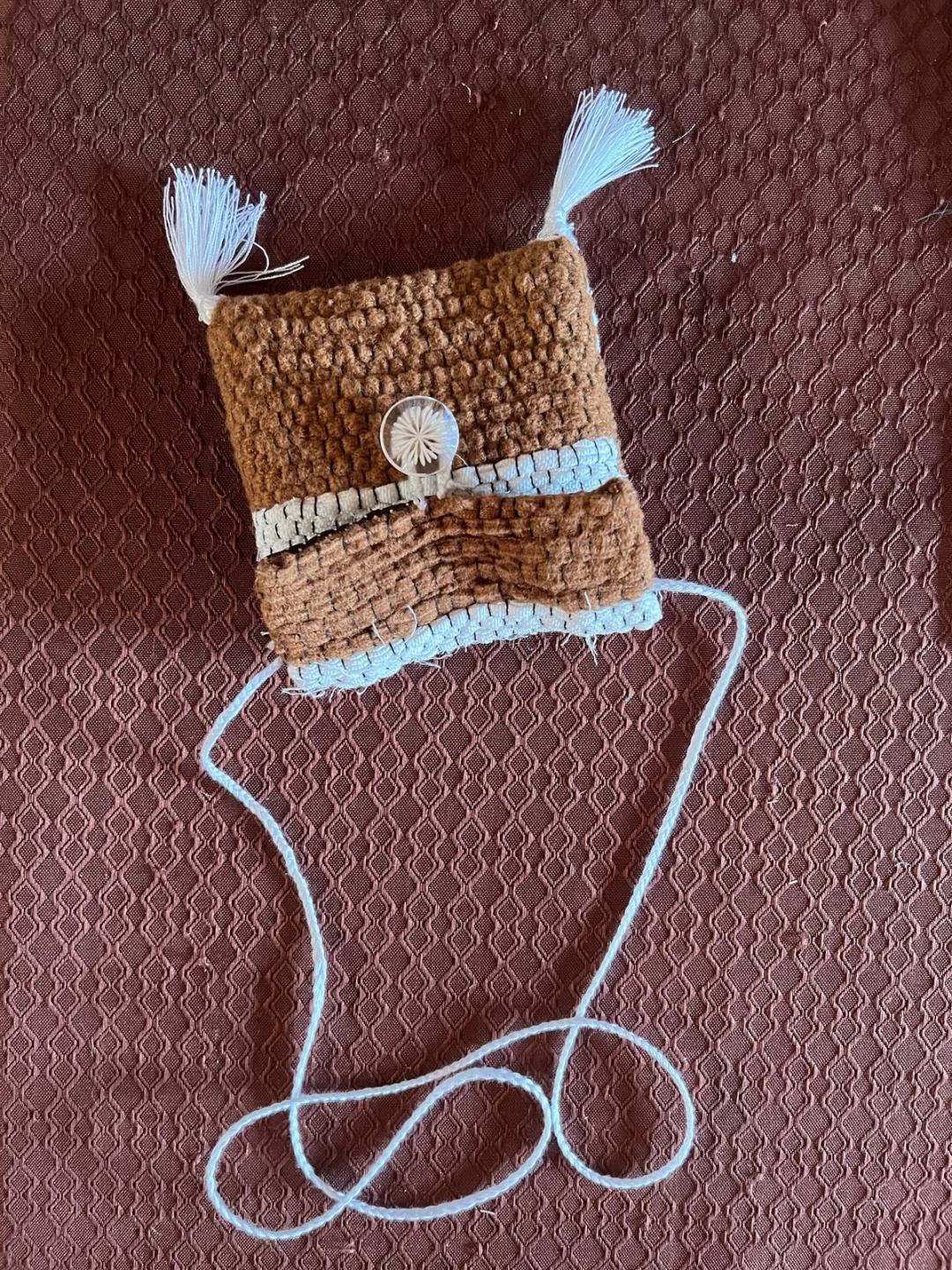 Pochette tissée marron et bleu clair, fermée par un bouton, avec deux pompons et une longue anse en fil.