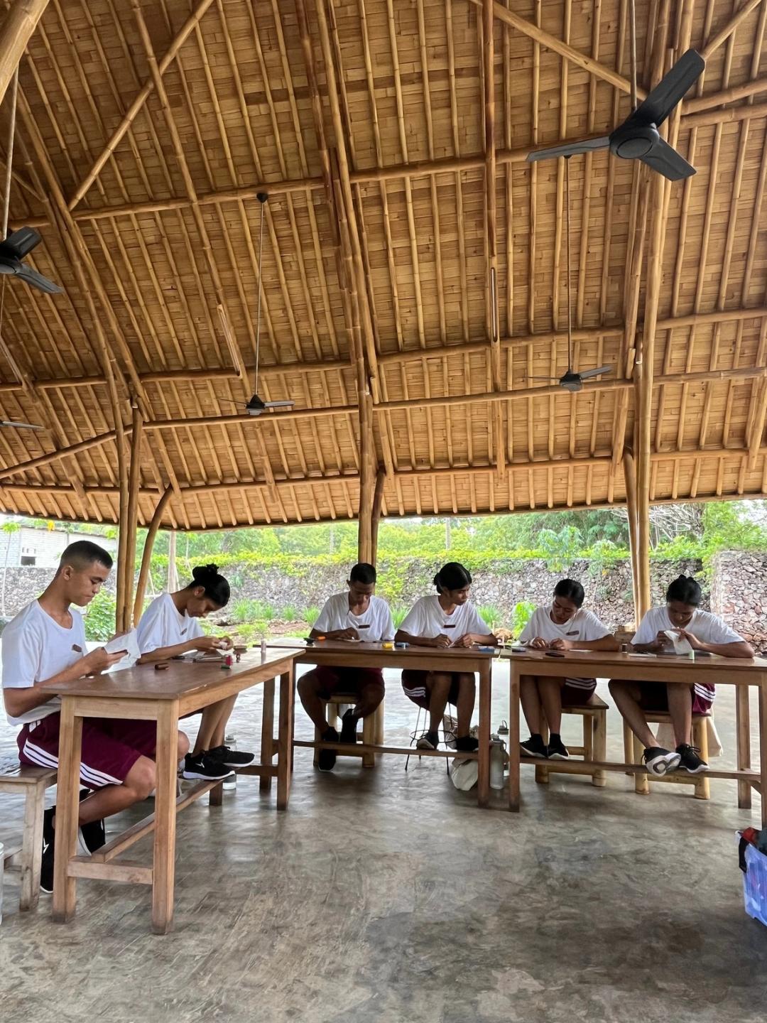 Élèves cousant à la main autour de tables en bois, sous un pavillon en bambou ouvert sur la végétation.