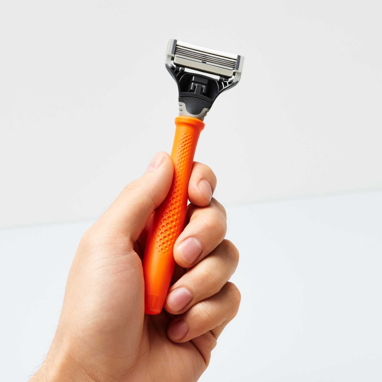 Hand holding an orange Truman razor, facing upward to show the five-blade cartridge and no-slip grip.