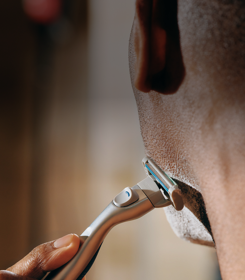 Man shaving his face with Harry's Plus razor against a blurred background