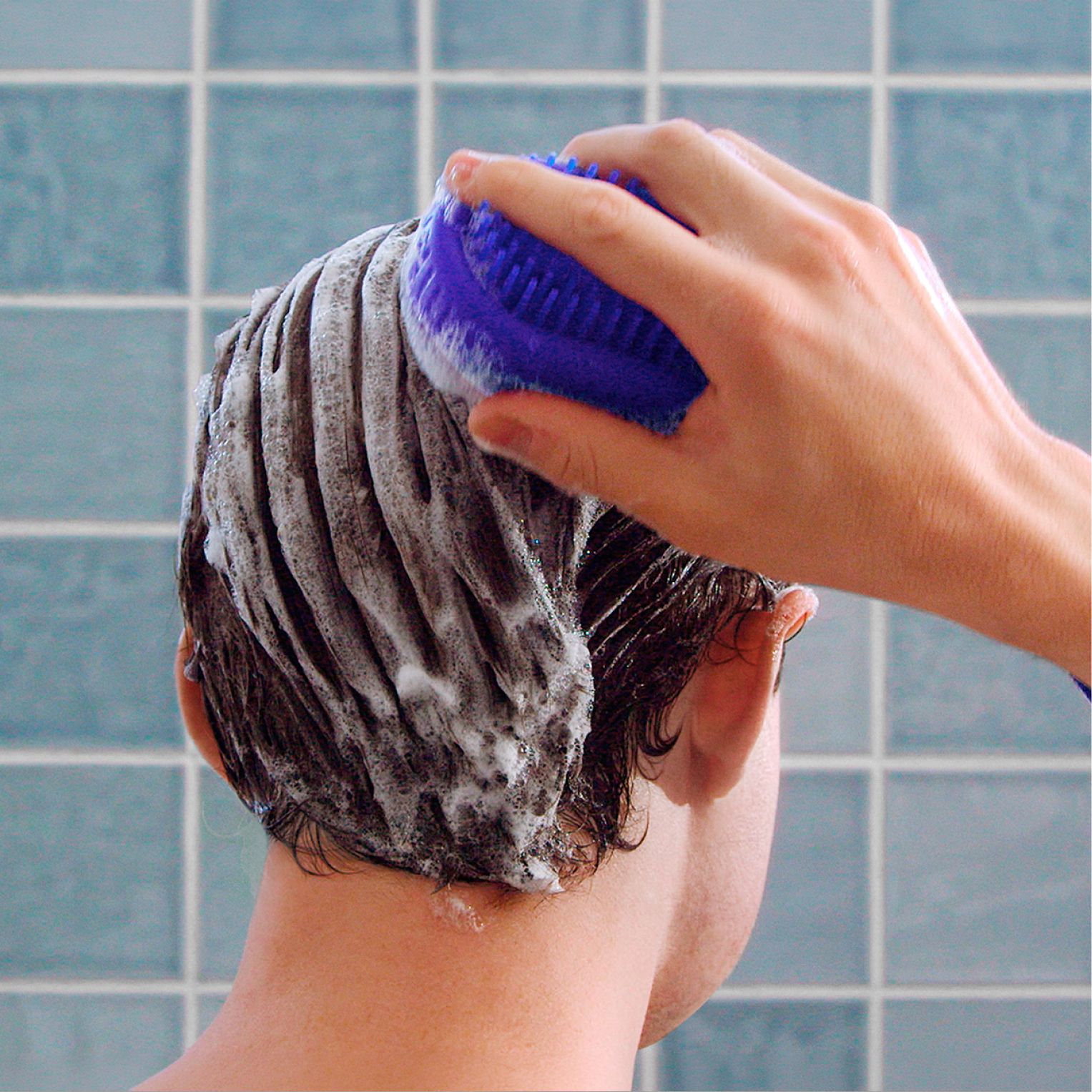 Blue scrubber being used on a soapy head of hair in blue tiled bathroom