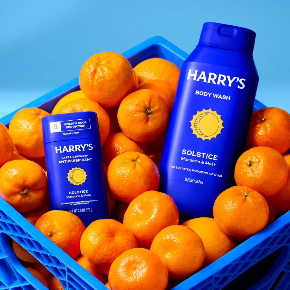 Two blue bottles with a picture of a yellow cartoon sun and Harry's logo in a blue crate of mandarin oranges against a lighter blue background.