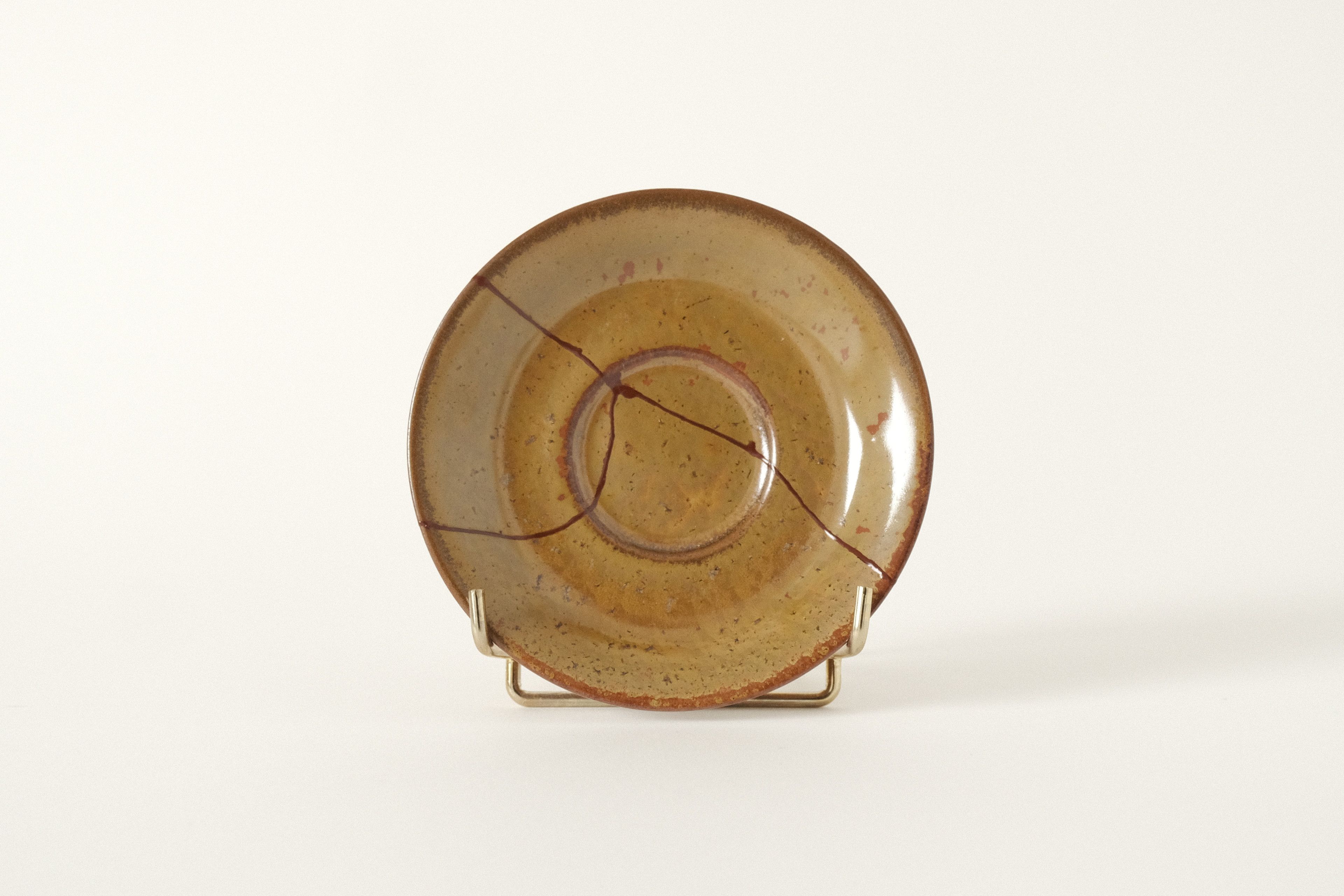 A small, brown ceramic saucer repaired with rust red kintsugi on an offwhite studio background.