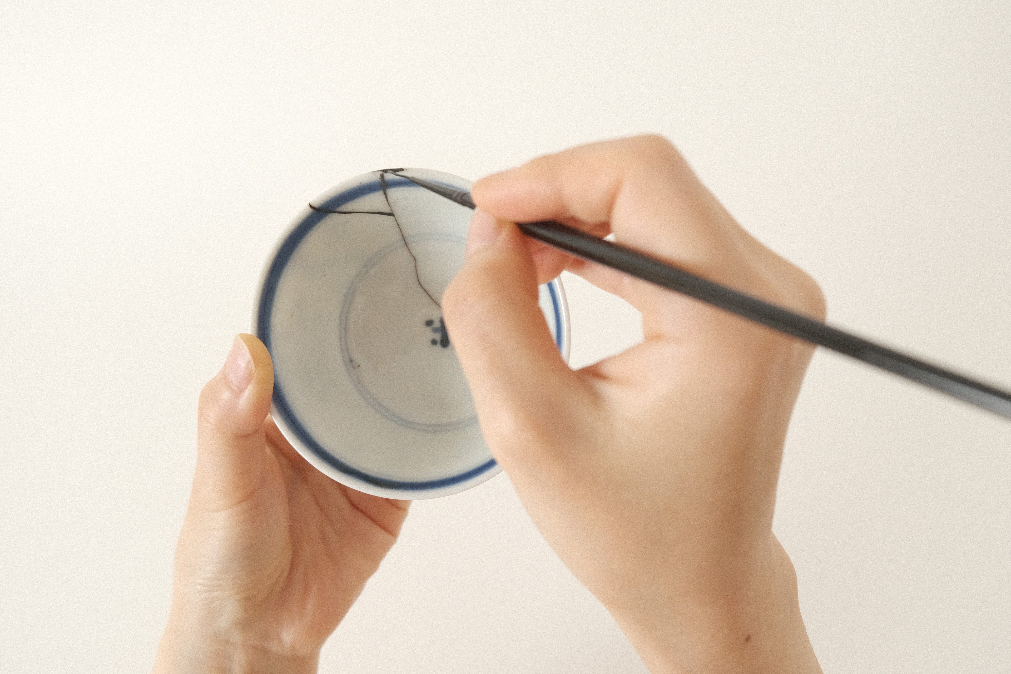 Hands holding a white porcelain bowl, applying finish to dark kintsugi cracks with a brush.
