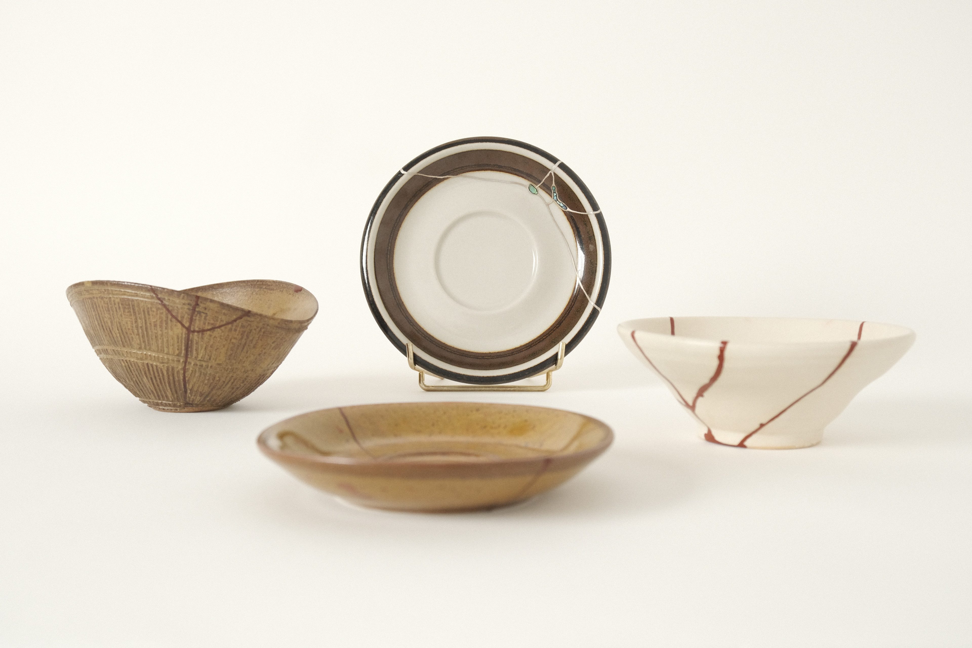 Four ceramic pieces with kintsugi seams on a pale studio background — a small bowl with red kintsugi seams at left, a dark-rimmed plate with tin kintsugi seams on a brass stand at centre, a low saucer in front with red kintsugi seams, and a brown bowl red kintsugi seams at right.