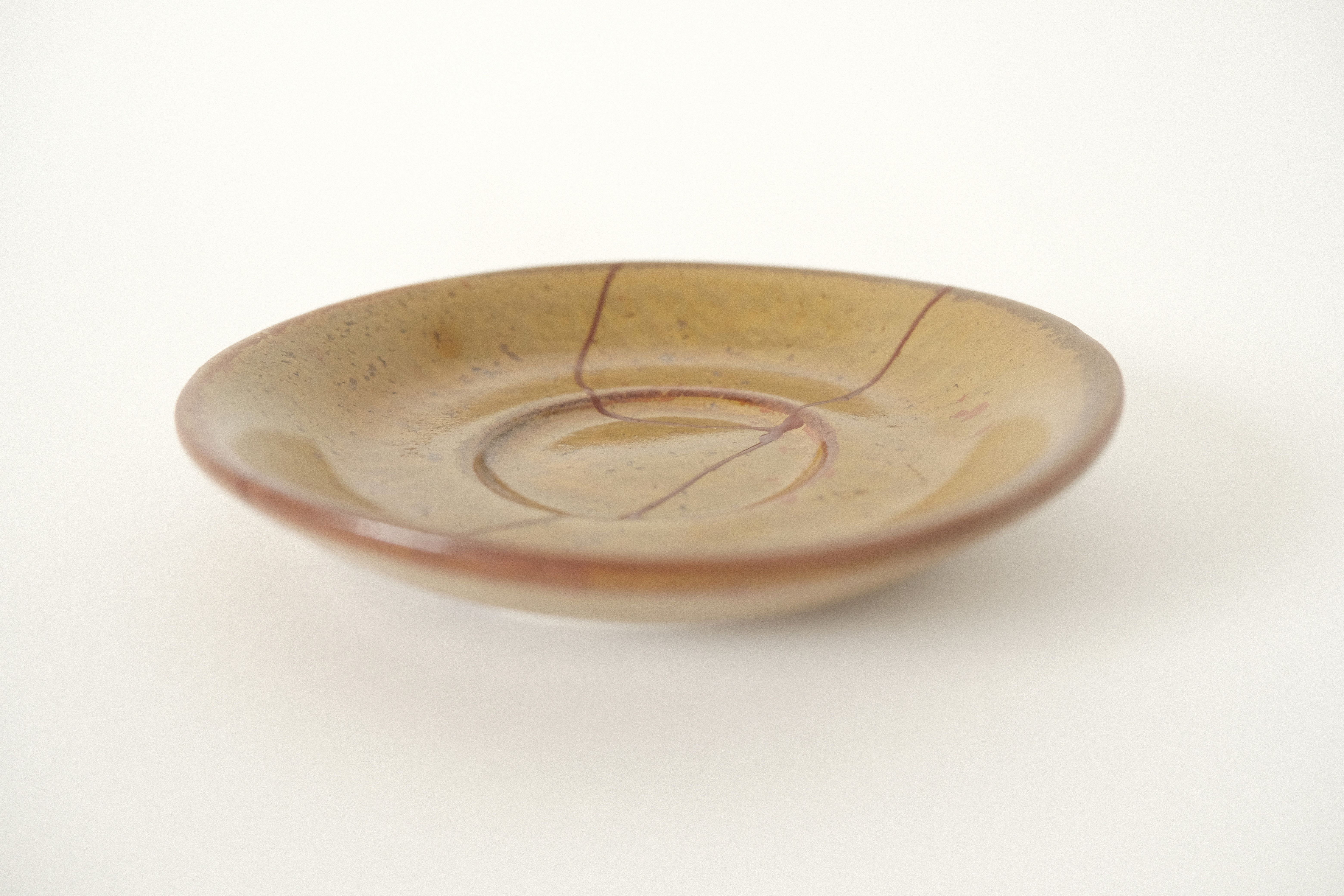 A small, brown ceramic saucer repaired with rust red kintsugi on an offwhite studio background.