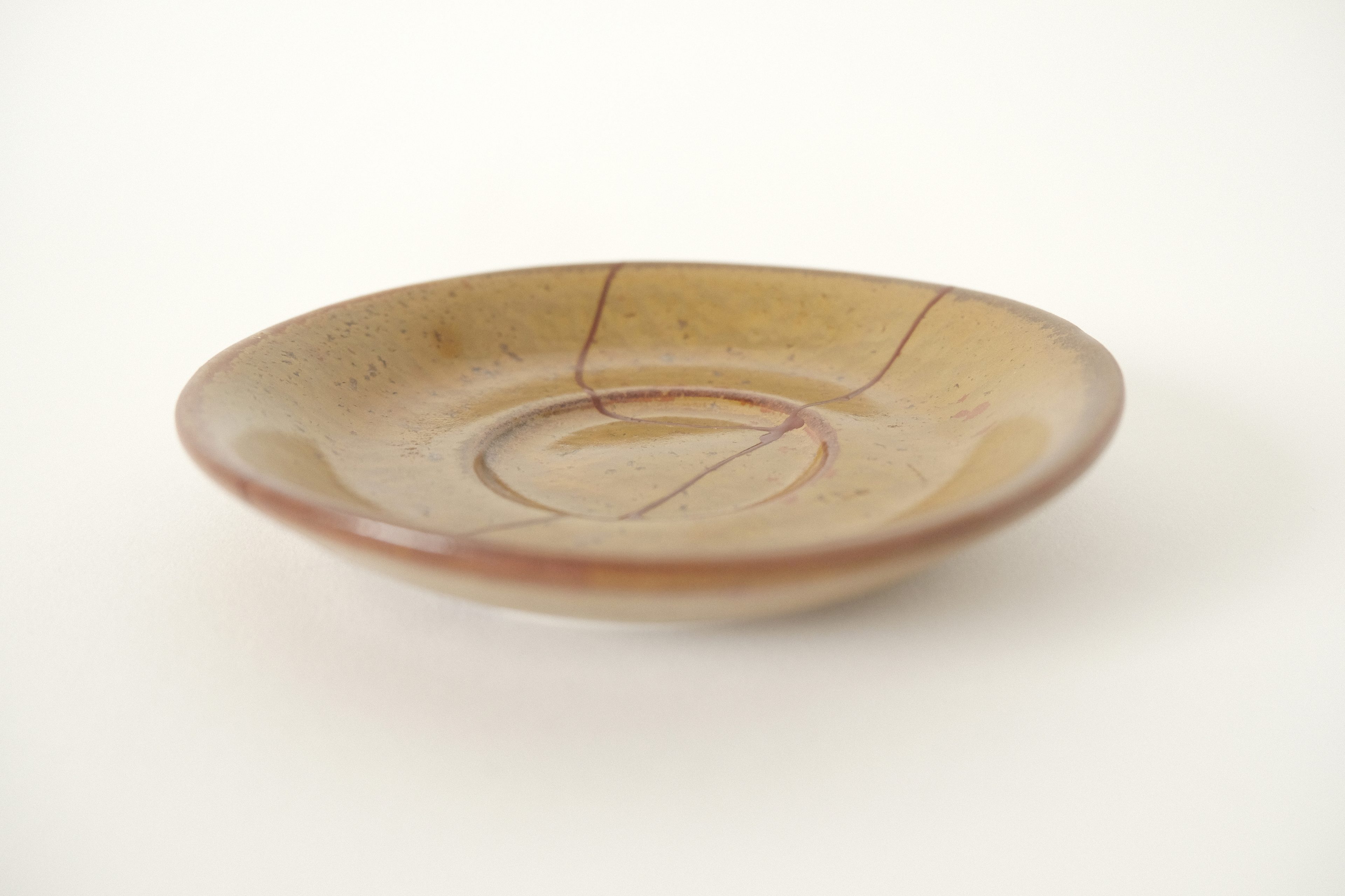 A small, brown ceramic saucer repaired with rust red kintsugi on an offwhite studio background.