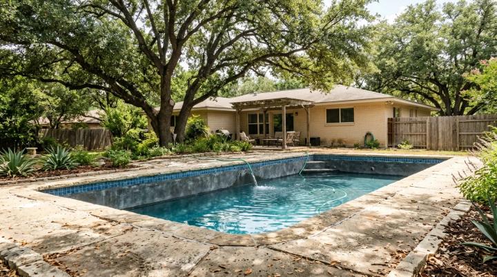 An eye-level afternoon shot of an in-ground gunite pool in an established Anderson Mill backyard (mature live oaks, limestone-edge decking, a single-story 1980s North Austin home in the background), with a clearly visible low water line a few inches below the tile and a garden hose topping it off. 