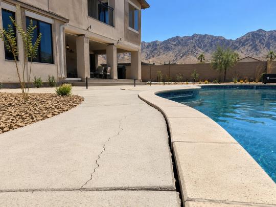 A clear, late-morning photo of a newer in-ground backyard pool behind a modern two-story stucco home in Enterprise, Las Vegas. Fresh desert landscaping with gravel and young plants.