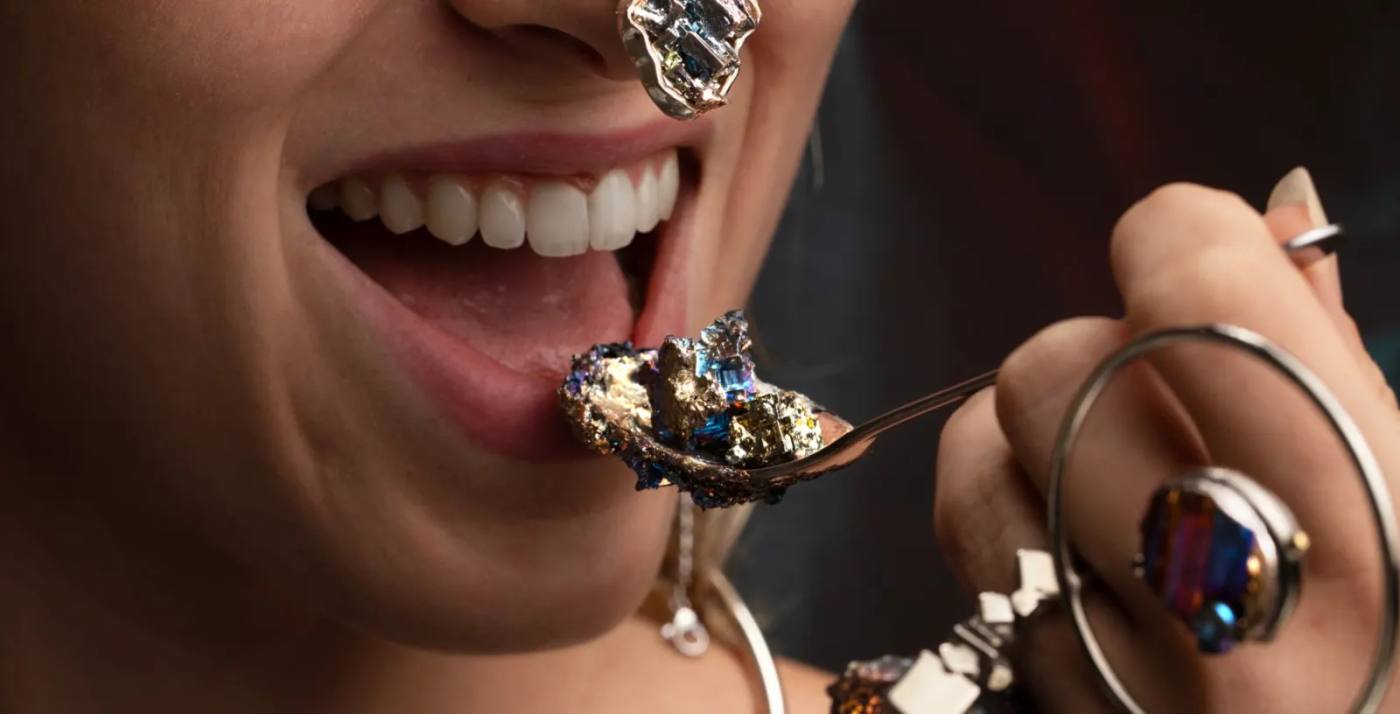 Bocca sorridente che accoglie da un cucchiaio una manciata di pietre e metalli preziosi.
