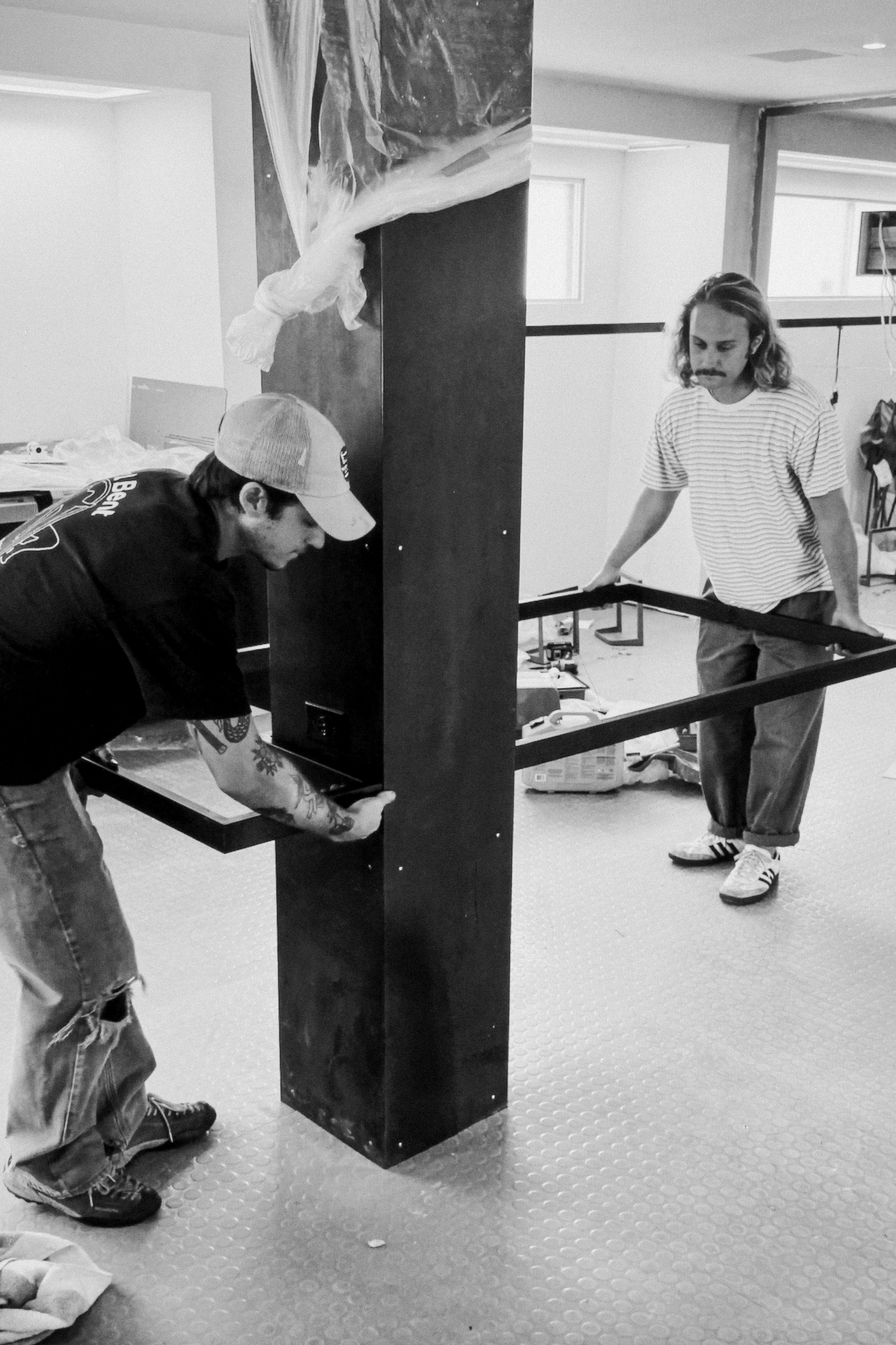 two men holding a steel desk frame against a steel column, WOE, Width Over Ears
