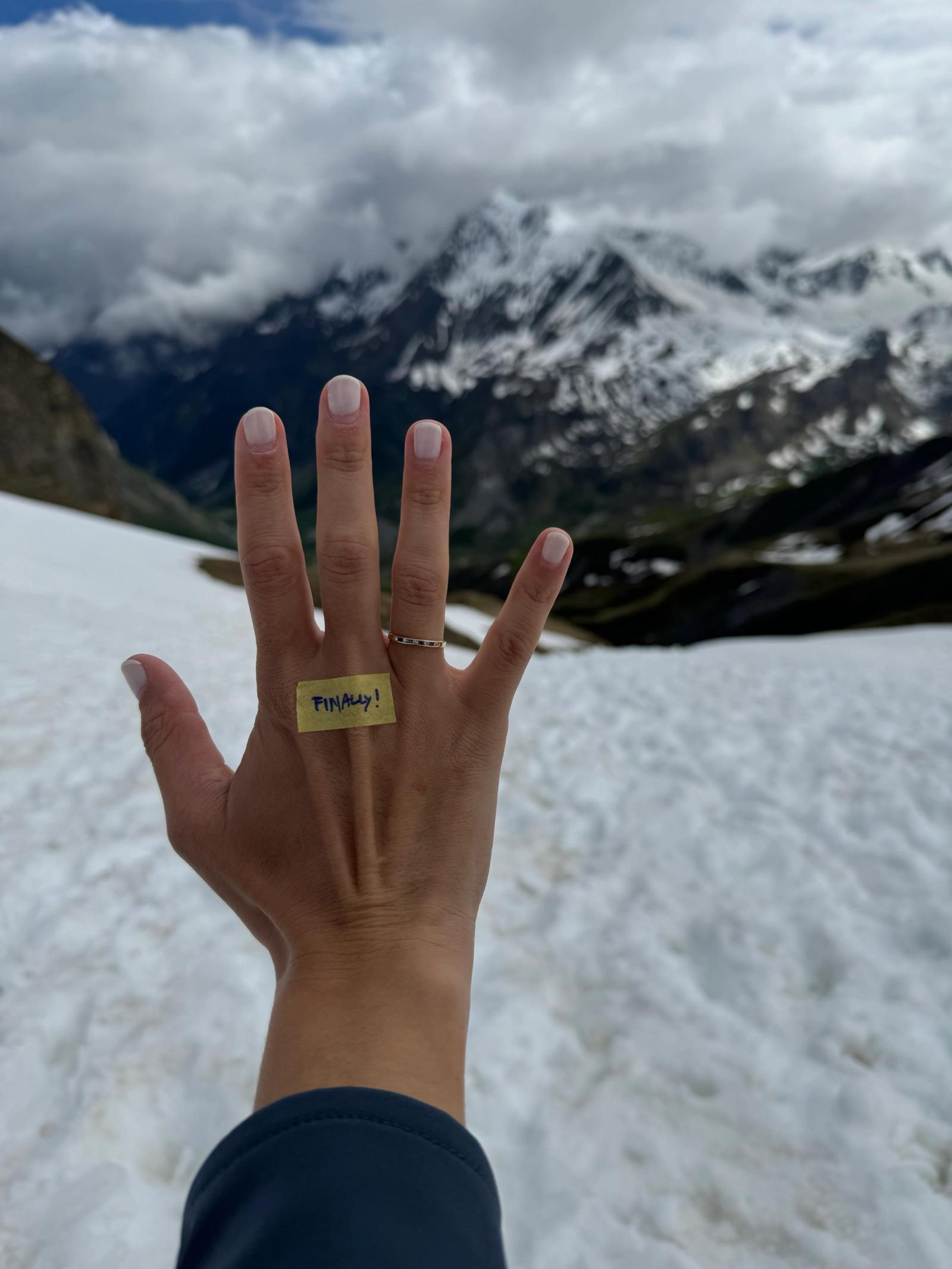 La mano de Flo con el anillo de compromiso y una nota que dice «FINALLY!», frente a montañas nevadas.