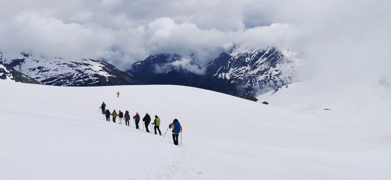 Un grupo de senderistas avanza en fila por la nieve, con montañas y nubes al fondo.