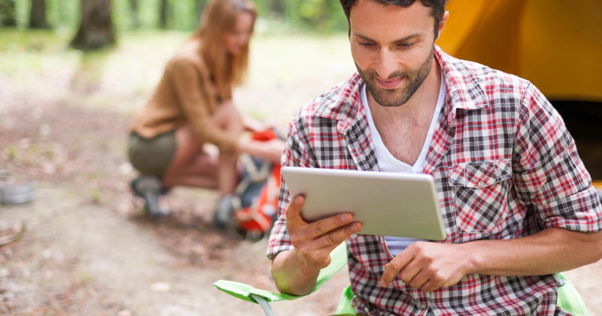 Man in plaid shirt using tablet at campground while woman sits by tent in forested camping area