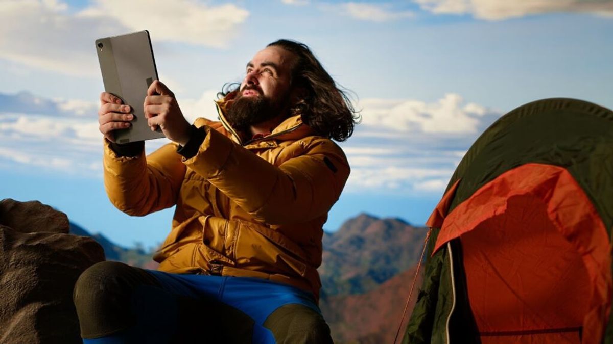 Bearded man in yellow jacket holding tablet while sitting outdoors near camping tent with mountainous landscape in background.