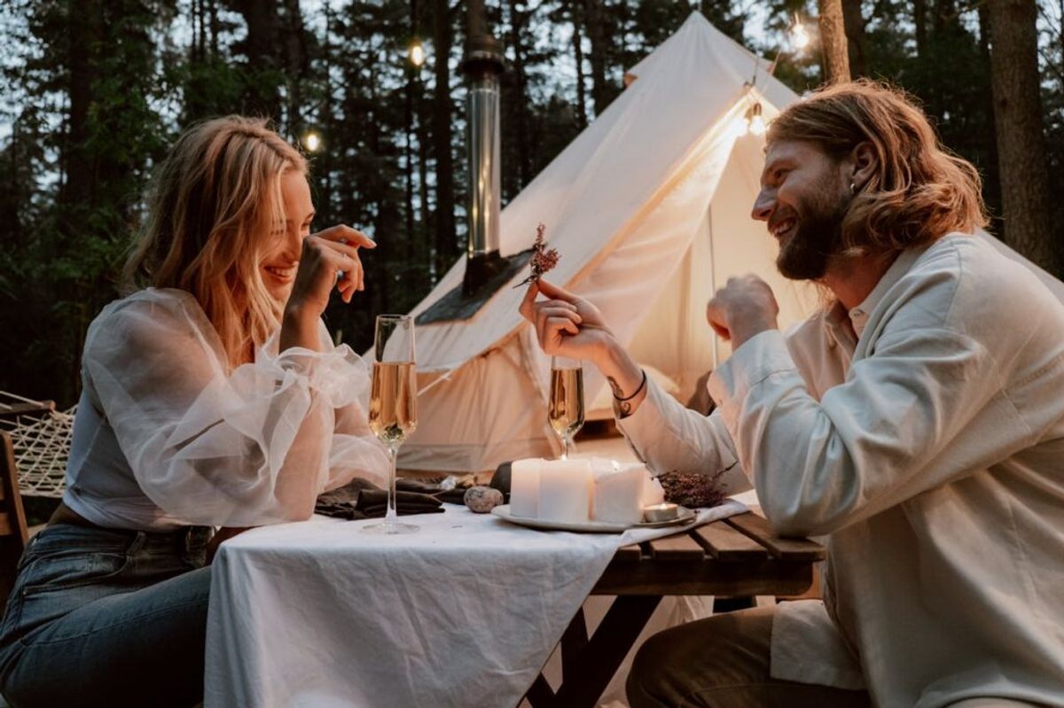 Couple enjoying candlelit dinner at outdoor table with champagne glasses in front of illuminated glamping tent surrounded by forest trees.