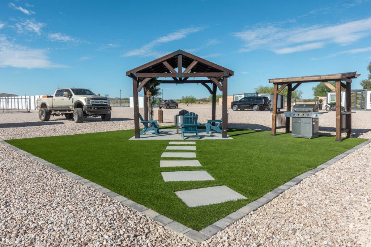 Modern RV resort site with artificial grass, wooden gazebo, teal chairs, outdoor kitchen, and RVs parked on gravel under blue sky