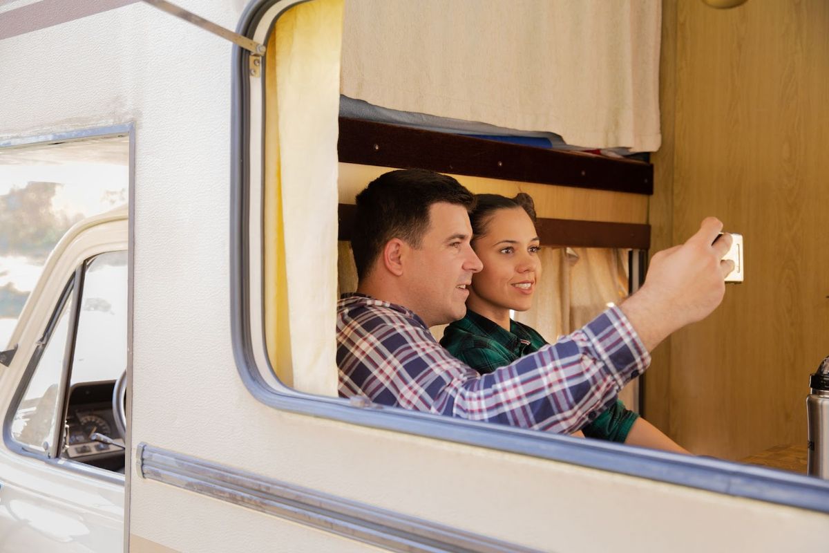 Couple looking out RV window at campsite, man pointing outside while woman smiles from inside motorhome interior.