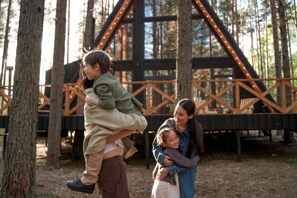 Family enjoying time outside a modern A-frame glamping cabin nestled in a pine forest, with string lights and wooden railings visible.