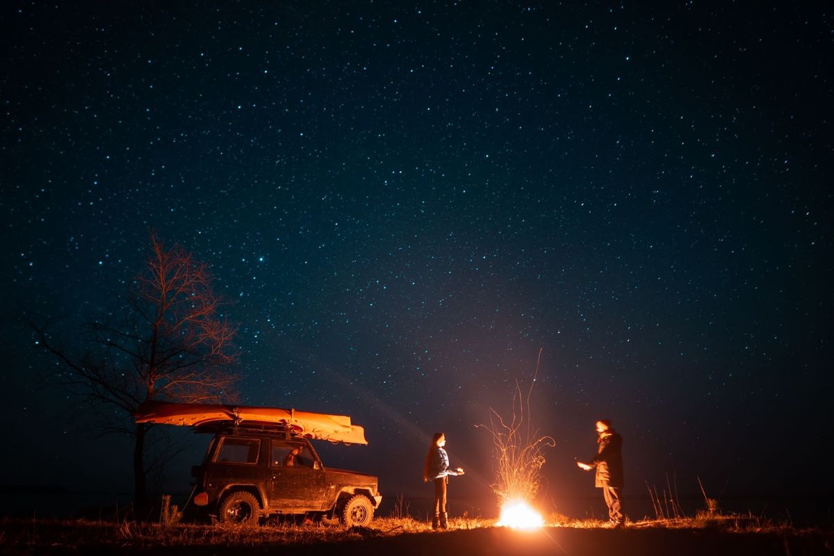 Two people around a glowing campfire next to a pickup truck under a starlit sky, representing outdoor hospitality and camping experiences.