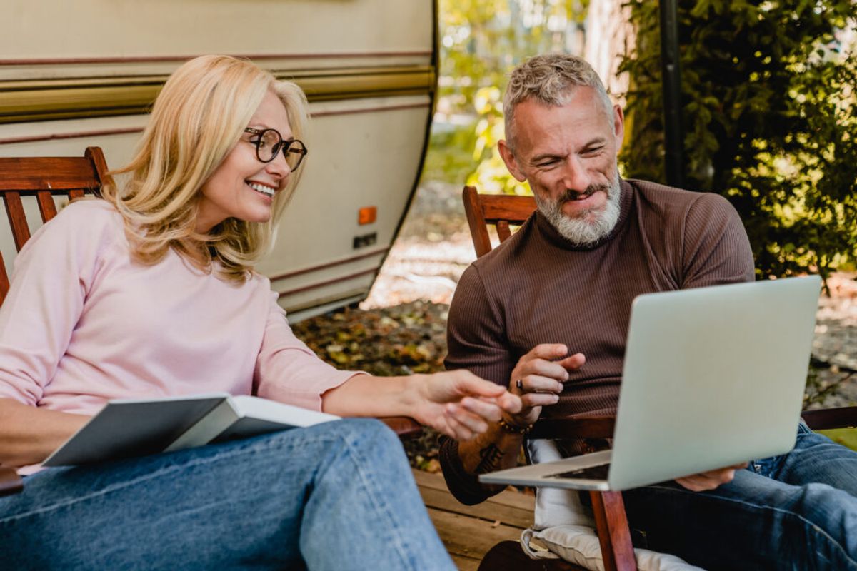 Two people sitting outdoors by an RV, smiling while reviewing documents and a laptop together at a wooden table.