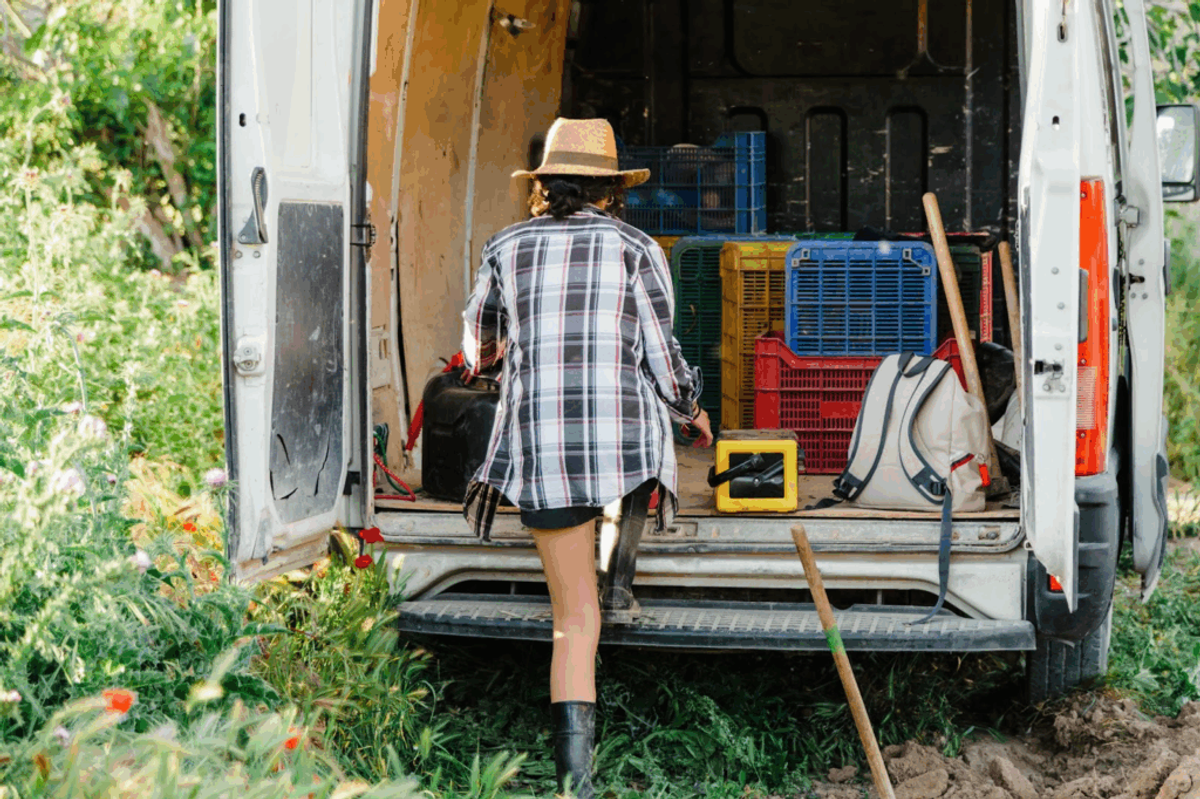 Worker in plaid shirt and straw hat organizing colorful plastic crates and tools inside white van, surrounded by green vegetation.