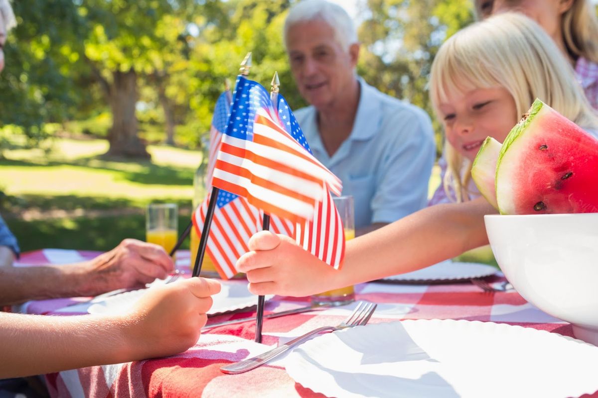 Family enjoying outdoor picnic with American flag, watermelon, and drinks at sunny park table.