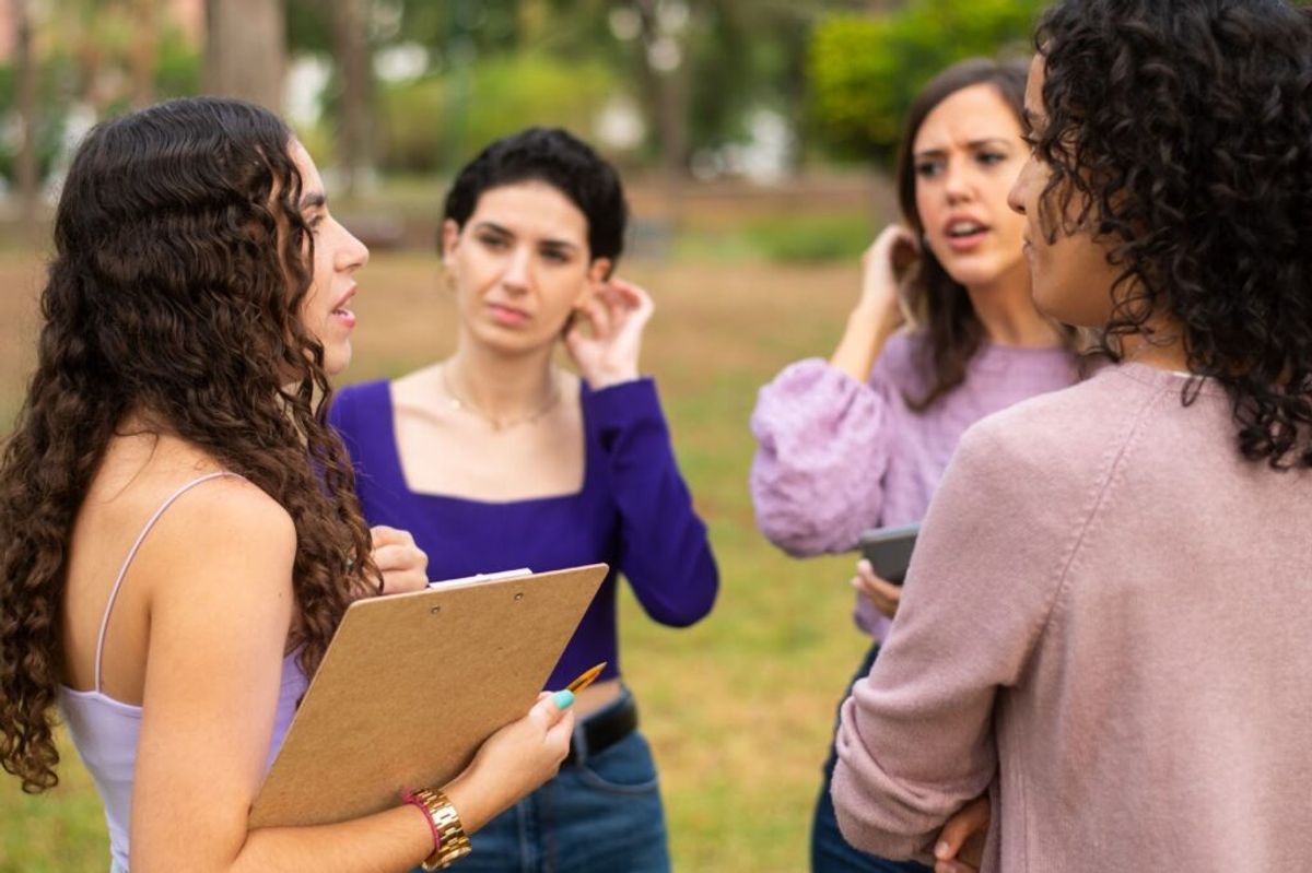 Group of women standing in a park having a discussion, one holding a clipboard, appearing to resolve a conflict or negotiate together outdoors.