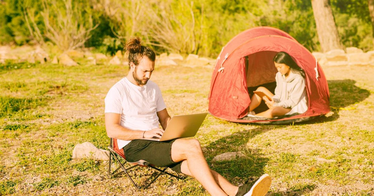 Man sitting on camping chair with laptop outside tent while woman works inside red tent at outdoor campsite.