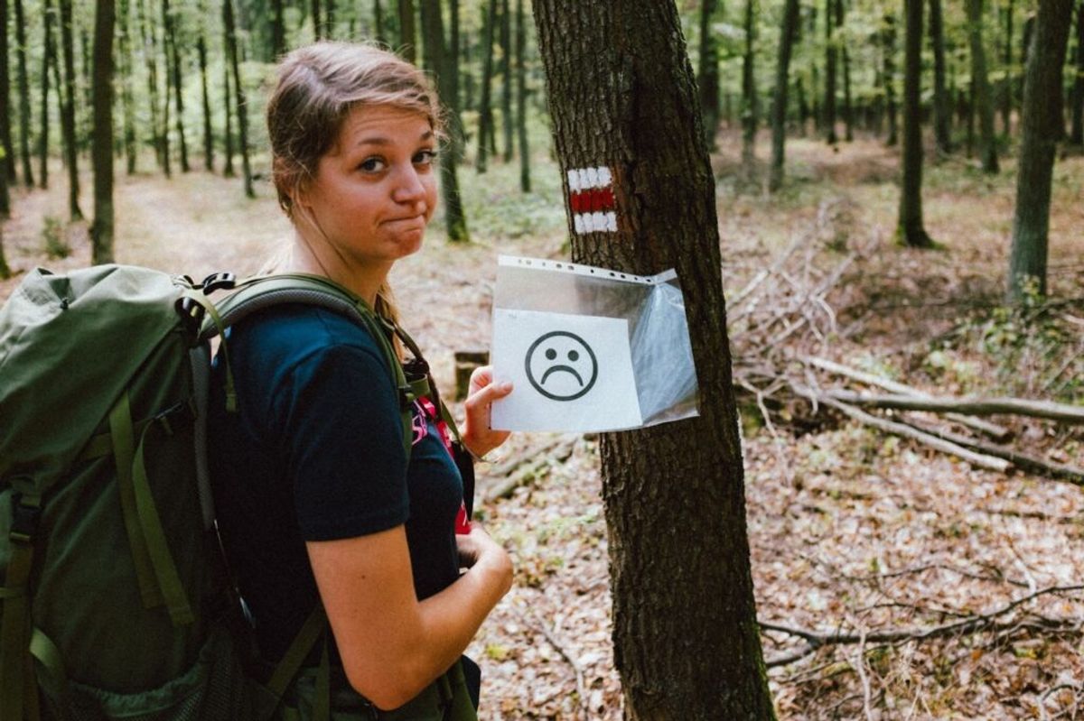 Hiker holding sad face sign on tree in forest, illustrating negative campground experience or rule violation.