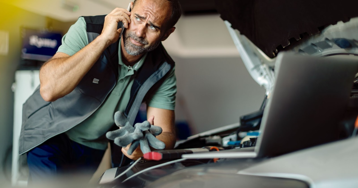 Mechanic on phone examining engine damage with tools and clipboard, discussing vehicle insurance claim.