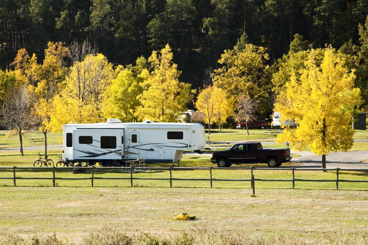 White travel trailer parked at a scenic campground with dark pickup truck, surrounded by golden aspen trees and wooden fence in autumn.