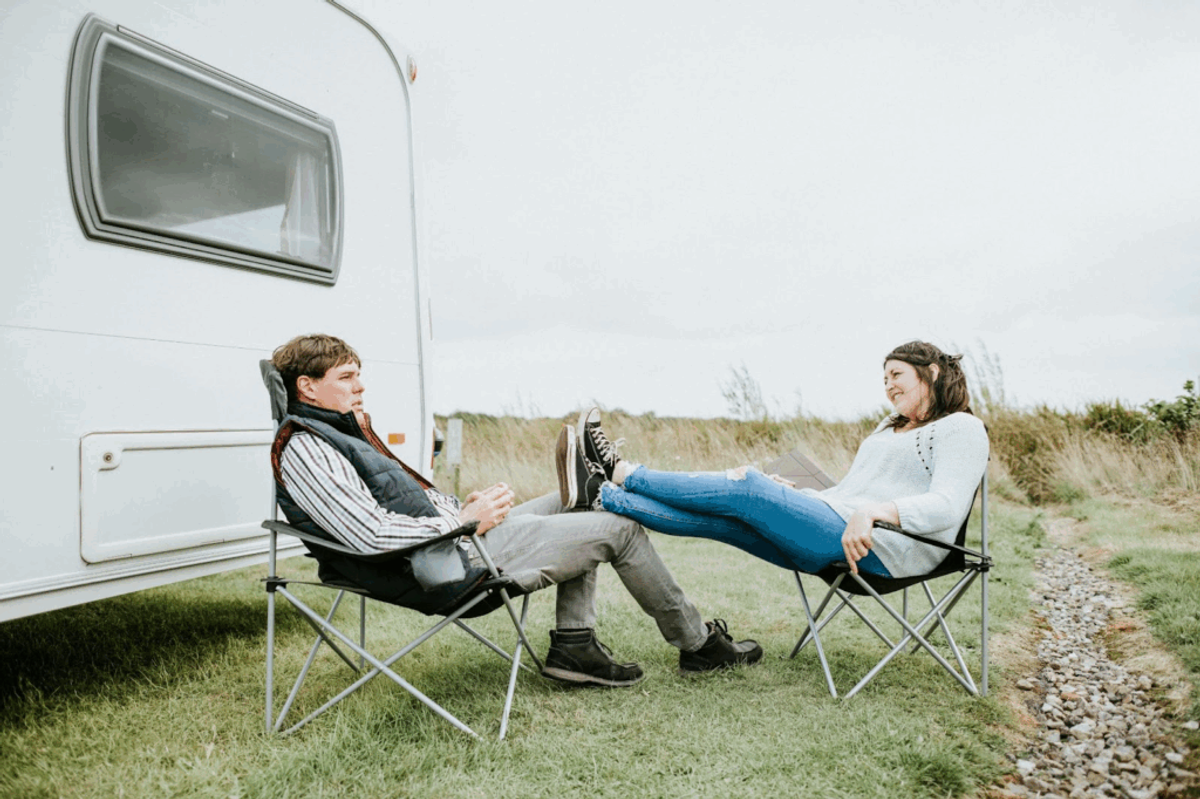 Two people sitting in camping chairs outside a white RV, relaxing in a grassy field with coastal vegetation in the background.