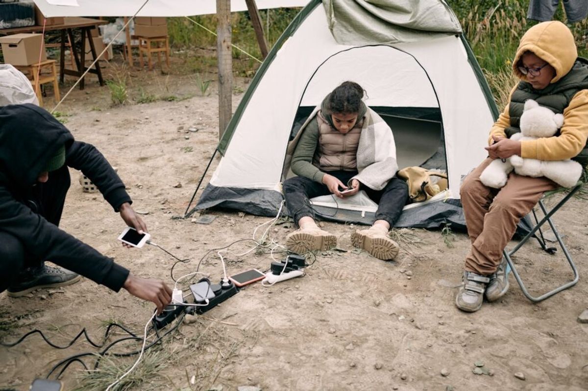 Three campers charging mobile devices at a tent campsite, demonstrating modern camping technology use outdoors.