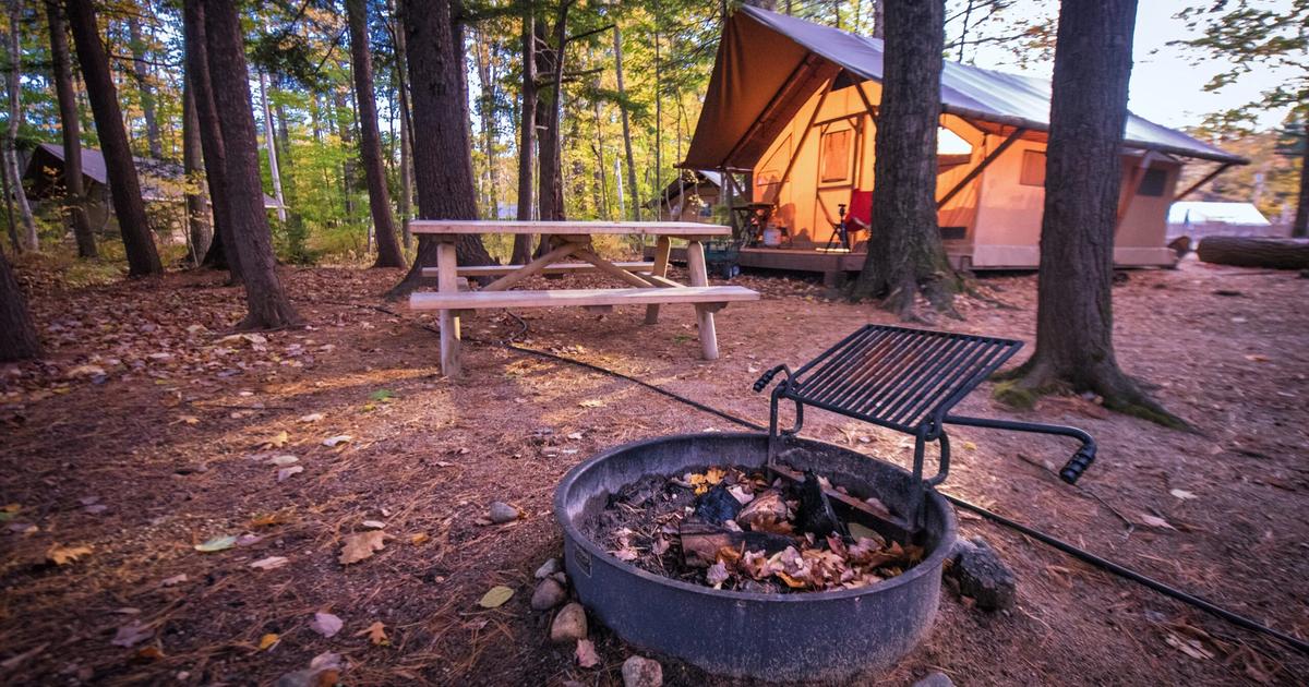 Glamping tent campsite with fire pit, grill, picnic table, and wooden benches surrounded by tall trees in autumn forest setting.