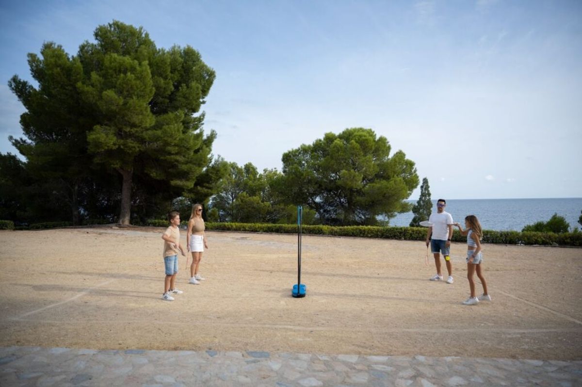 Families playing volleyball on a sandy beach with pine trees and calm sea in the background at a seaside campsite.