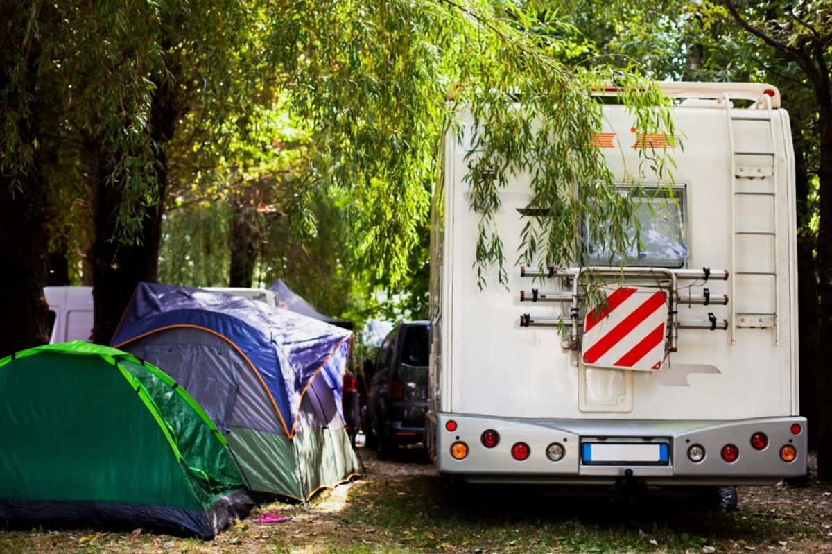 White RV parked at campsite with green and blue tent nearby, surrounded by trees and camping gear, showing typical campground setup.