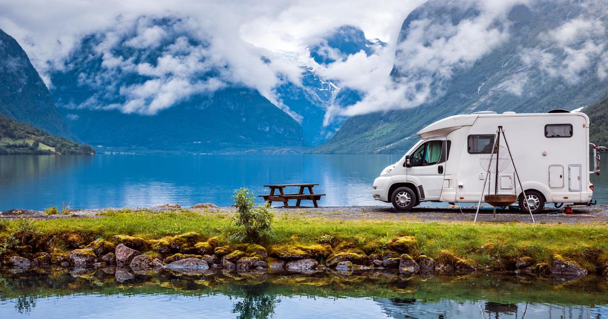A white camper van in front of a lake with their reflection in the water with a few mountains