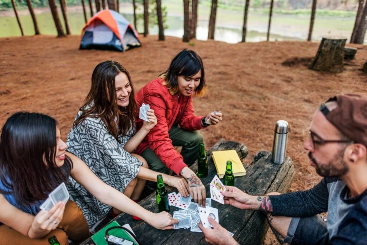 Group of friends playing cards at a wooden log table at a forest campsite with a tent and water visible in the background.