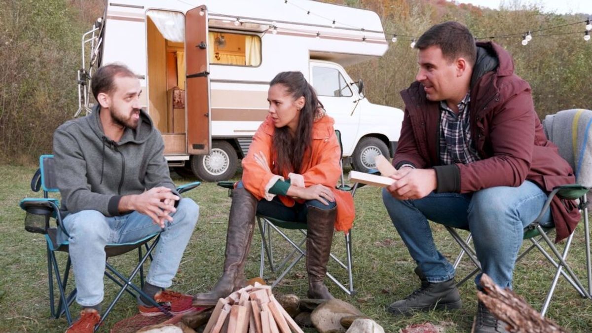 Three people sitting in camping chairs around a fire pit next to a vintage RV, having a discussion in an outdoor campsite setting.