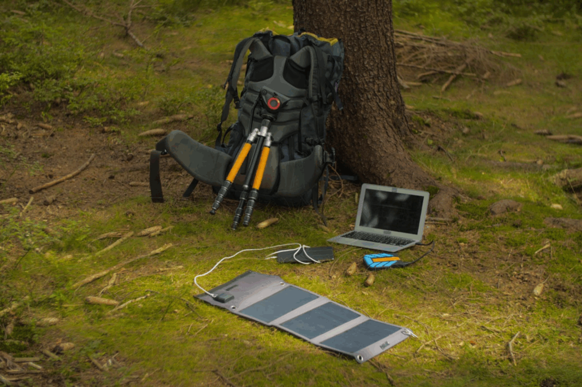 Portable solar panel charging a laptop and smartphone in a forest campsite next to a backpack and hiking equipment.