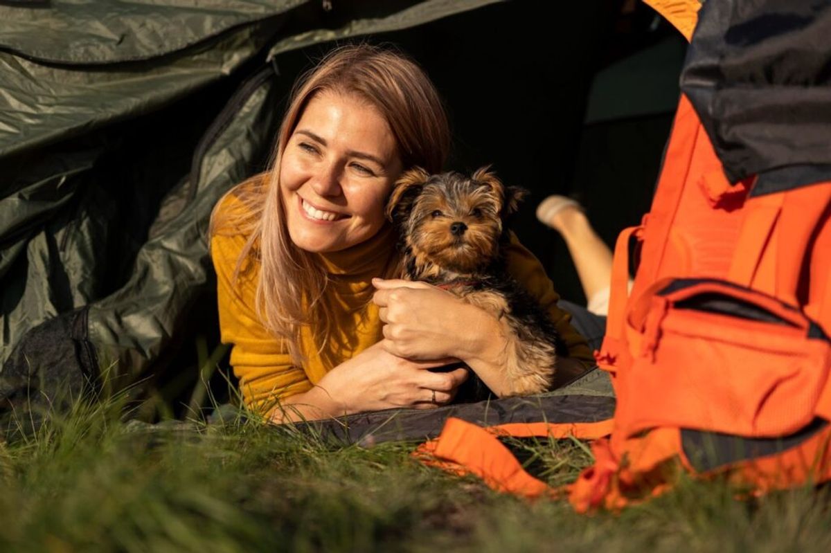 Woman holding a small dog inside a tent wearing orange life vest, camping outdoors.