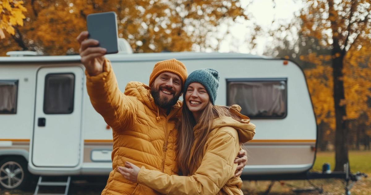 Happy couple in winter coats taking a selfie in front of their camper trailer during autumn, surrounded by golden trees.
