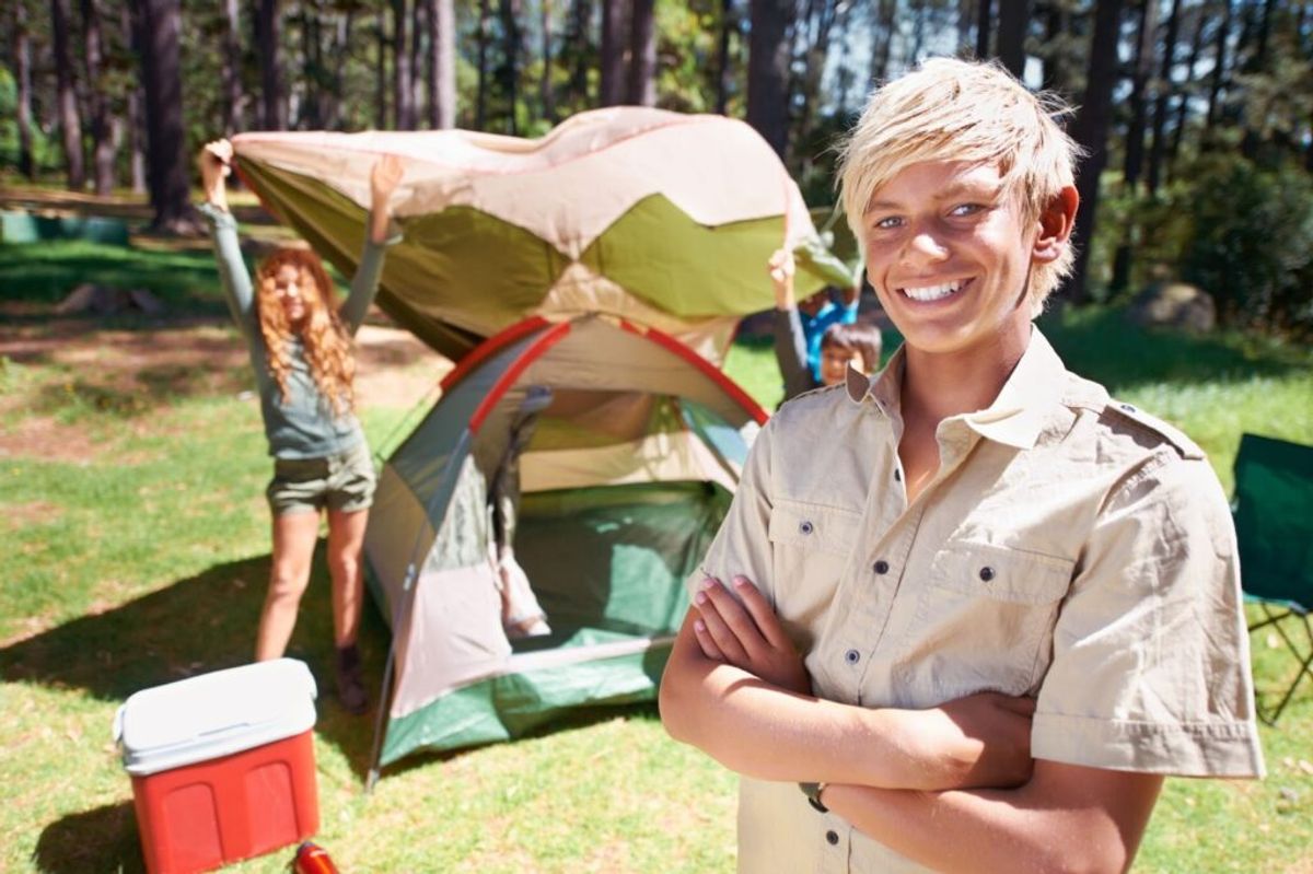 Young boy in khaki shirt smiling at camera near tent in forest campground with child holding rope in background.