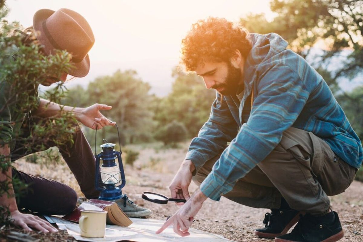 Two campers studying a map together outdoors at sunset, with a lantern and mug on the ground beside them.