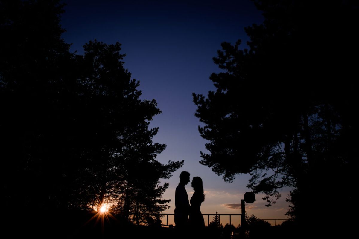 Silhouetted couple embracing at sunset on a railing overlooking trees and sky, with a basketball hoop visible nearby.